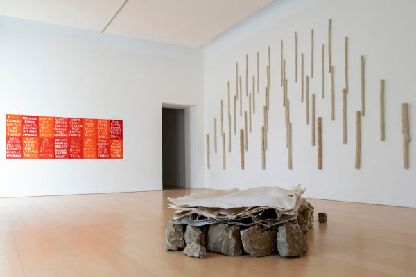 Museum wall with driftwood arranged to fake perspective, rocks in the center of the room covered in cloth, red and orange painted rectangles with white inscription on left wall.jpg